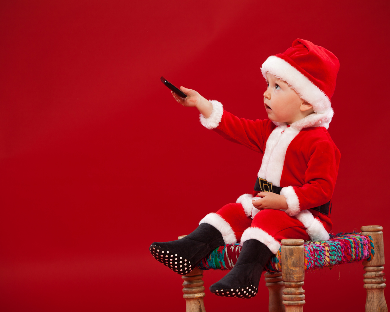 Маленький мальчик в костюме Санта Клауса сидит на стуле на красном фоне