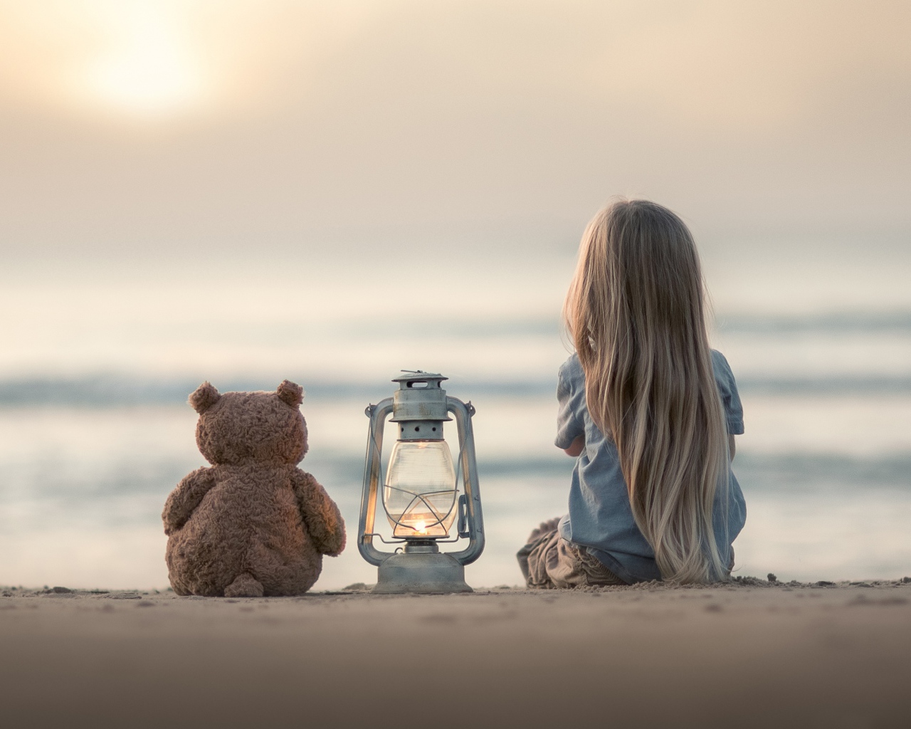 Маленькая девочка сидит на песке с плюшевым мишкой 