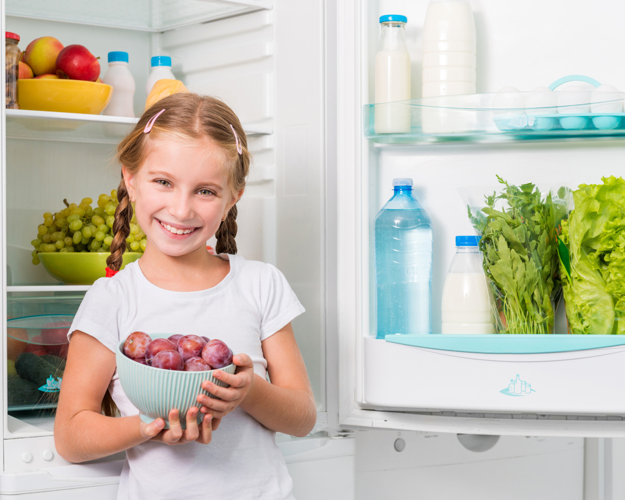 Маленькая девочка с миской со сливами в руках у холодильника 
