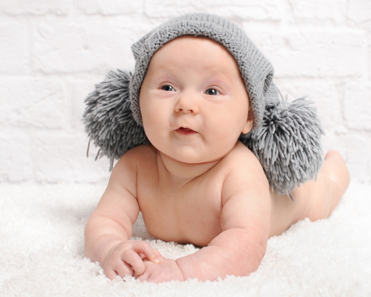 Улыбающийся грудной ребенок в серой шапке с бубонами