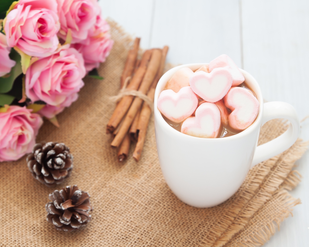 Чашка какао с маршмеллоу на столе с корицей, шишками и букетом розовых роз