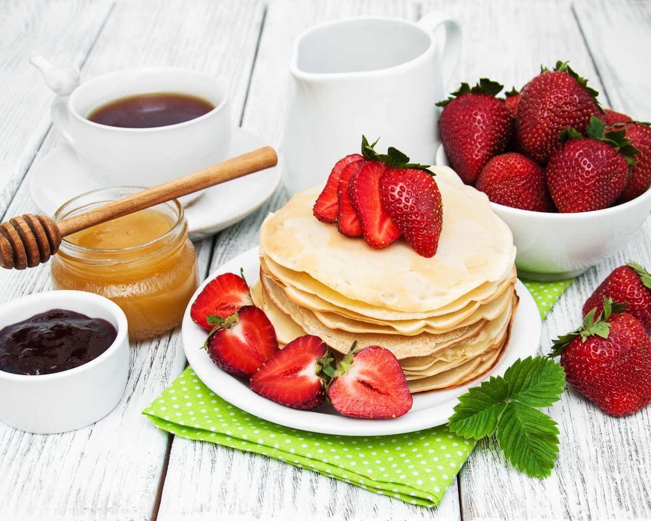 Тонкие блины на столе с ягодами клубники, медом, чаем и джемом