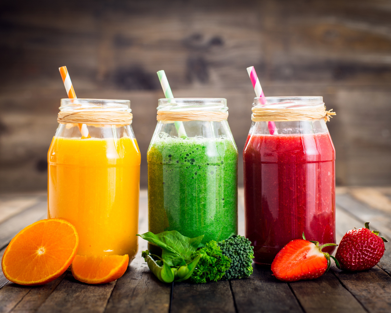 Три банки смузи с трубочкой на столе с апельсином, брокколи и клубникой