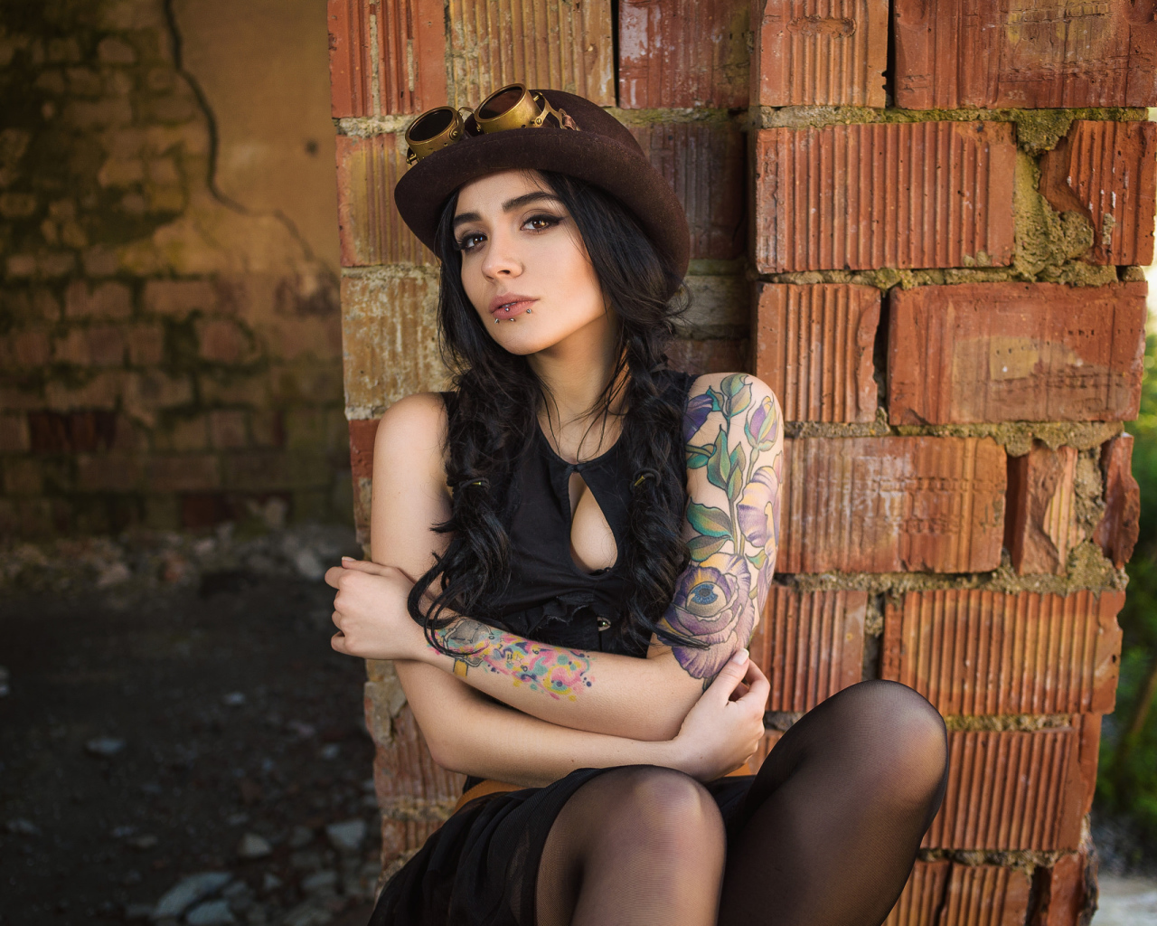 Девушка в шляпе с татуировками на руке сидит у стены