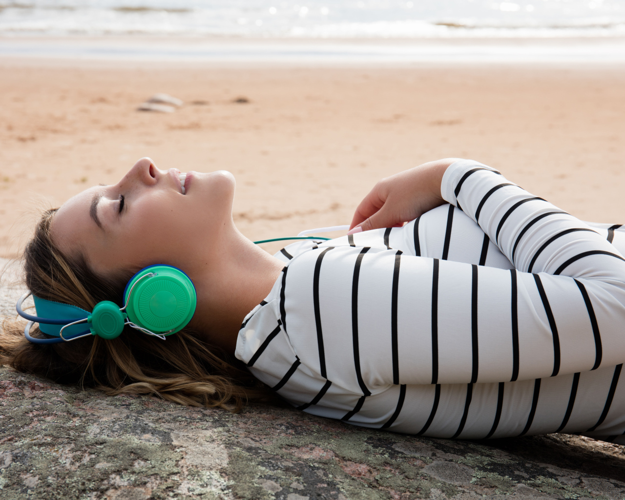 Молодая девушка лежит в наушниках на земле у моря
