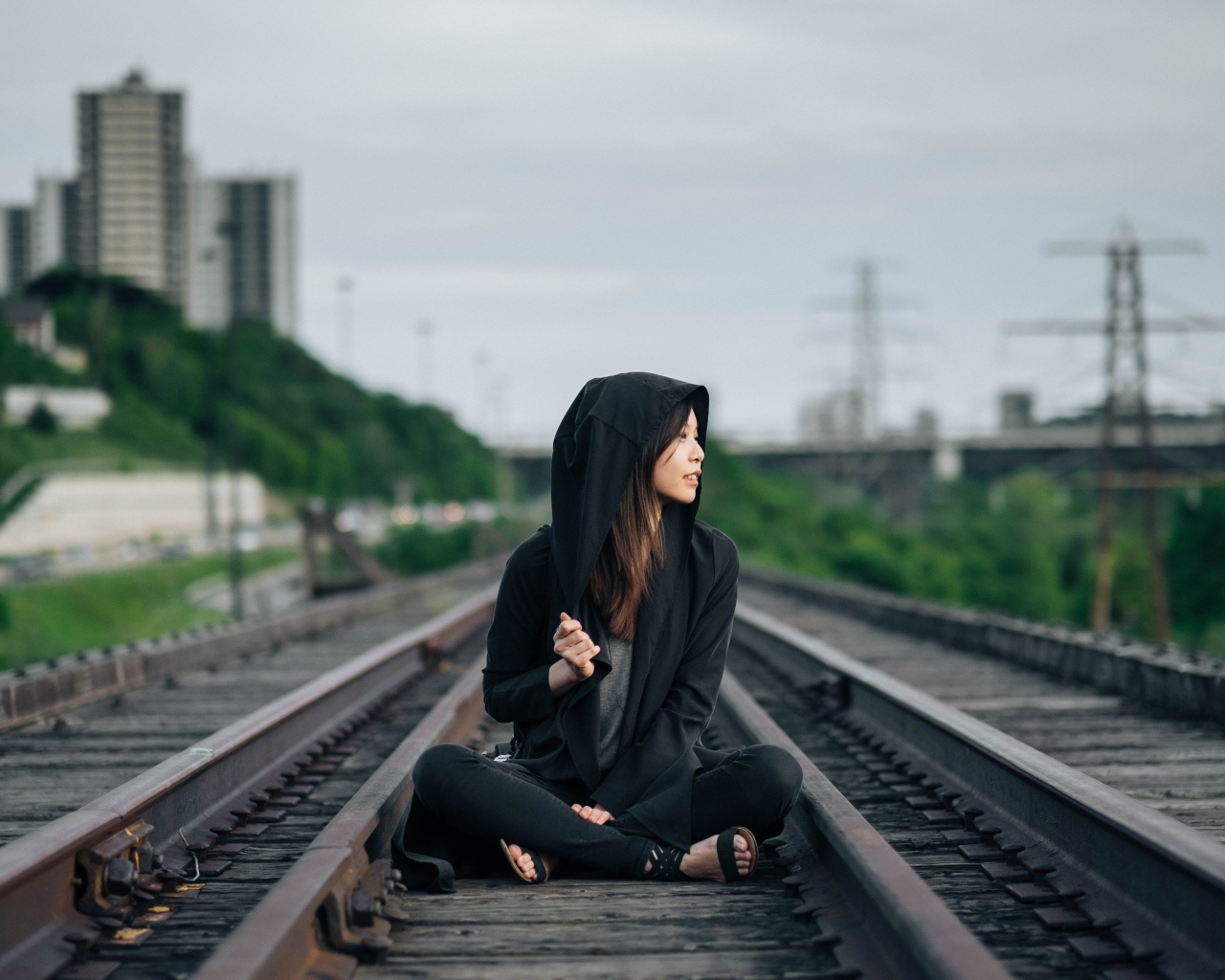 Девушка азиатка в черном балахоне сидит на рельсах 