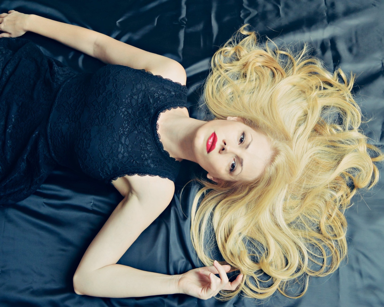 Длинноволосая блондинка в черном платье лежит на кровати