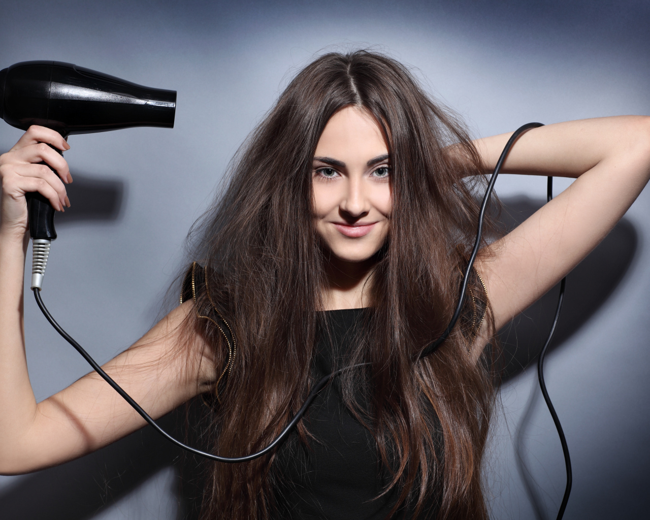 Длинноволосая девушка сушит волосы феном на сером фоне