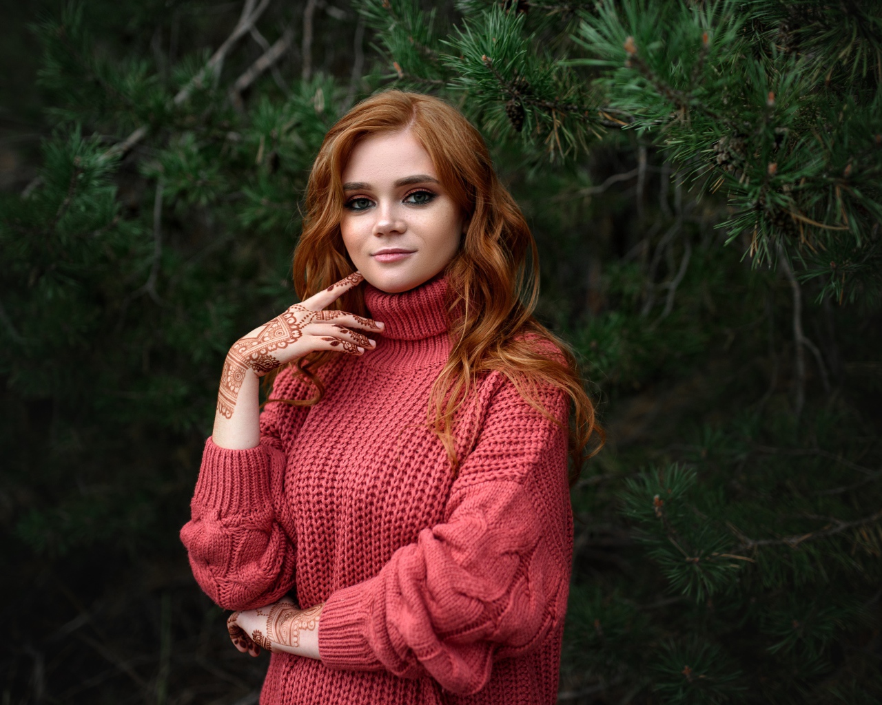 Рыжеволосая девушка в розовом свитере стоит у ели