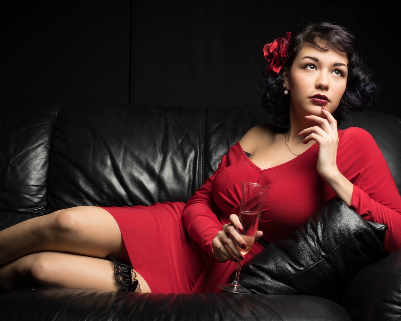 Девушка в красном платье с бокалом в руке лежит на кожаном диване