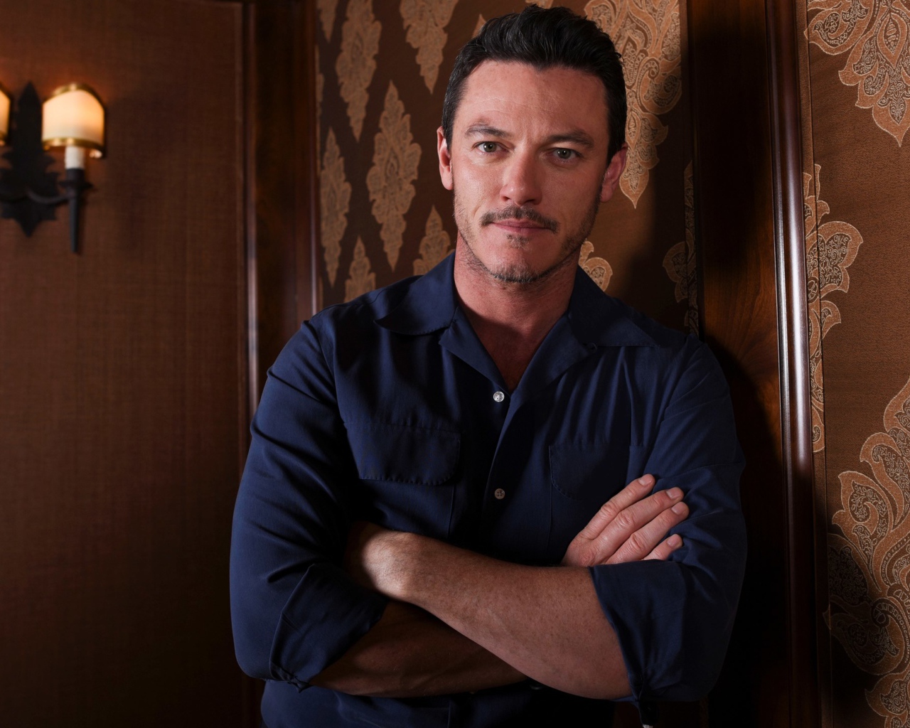 Актер Люк Эванс в рубашке стоит у стены