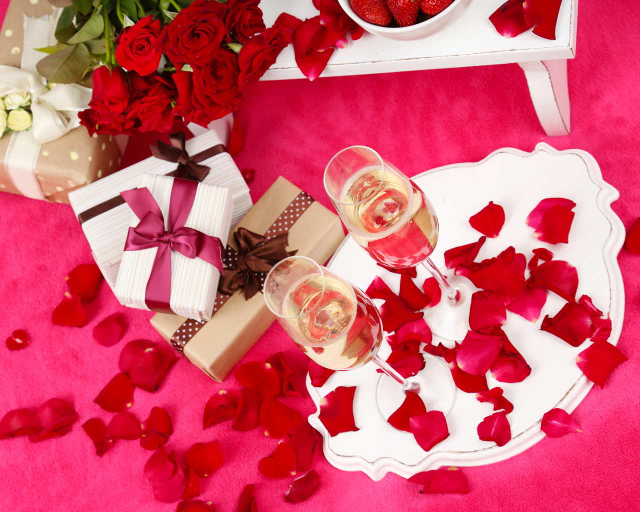 Два фужера с шампанским на красном покрывале с лепестками, подарками и букетом роз для любимой