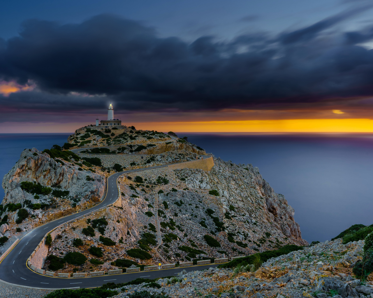 A winding road leads to a lighthouse on the edge of a cliff under a cloudy sky