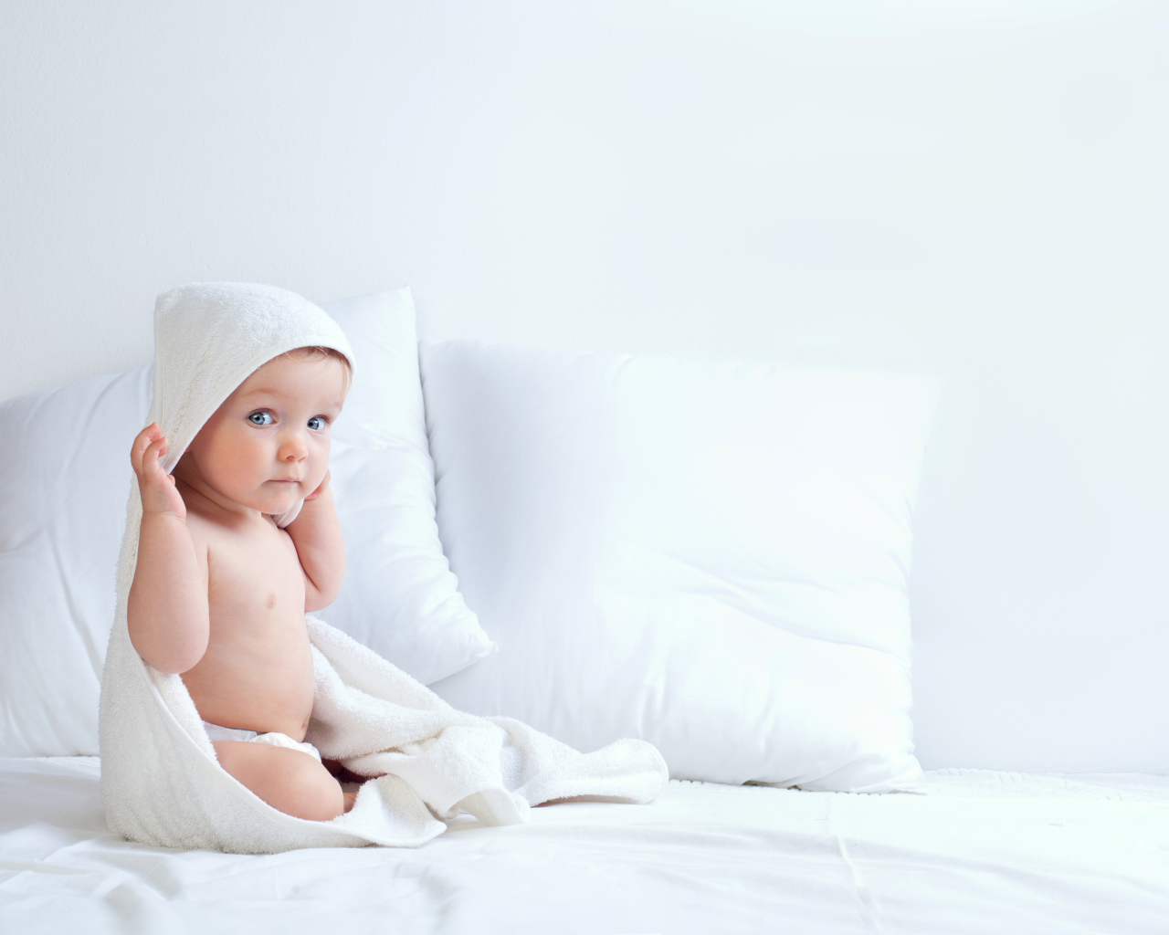 Маленький мальчик сидит в белом покрывале на кровати с подушками