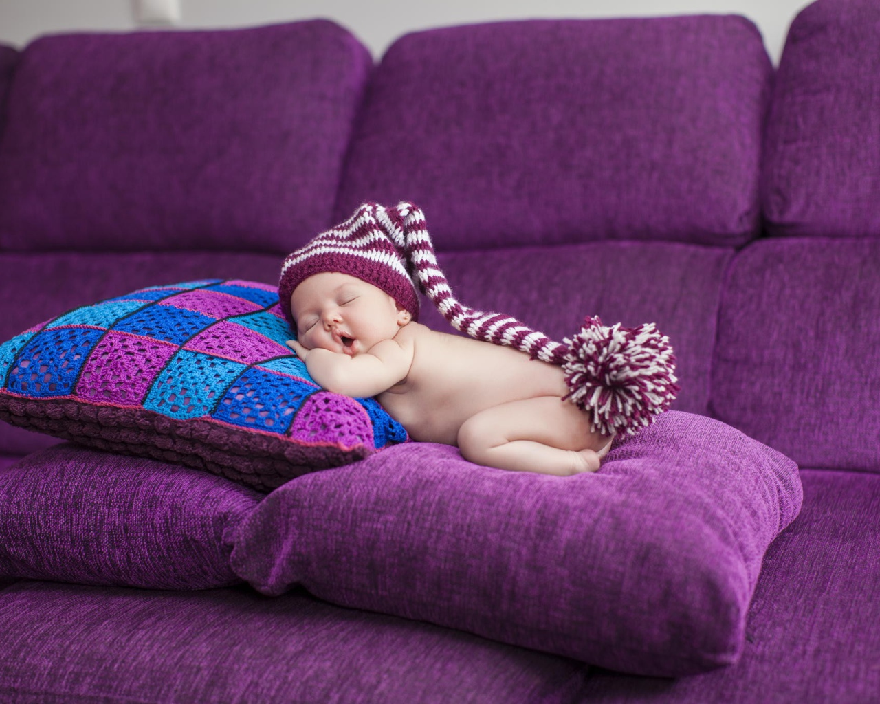 Маленький ребенок в большой шапке спит на фиолетовых подушках