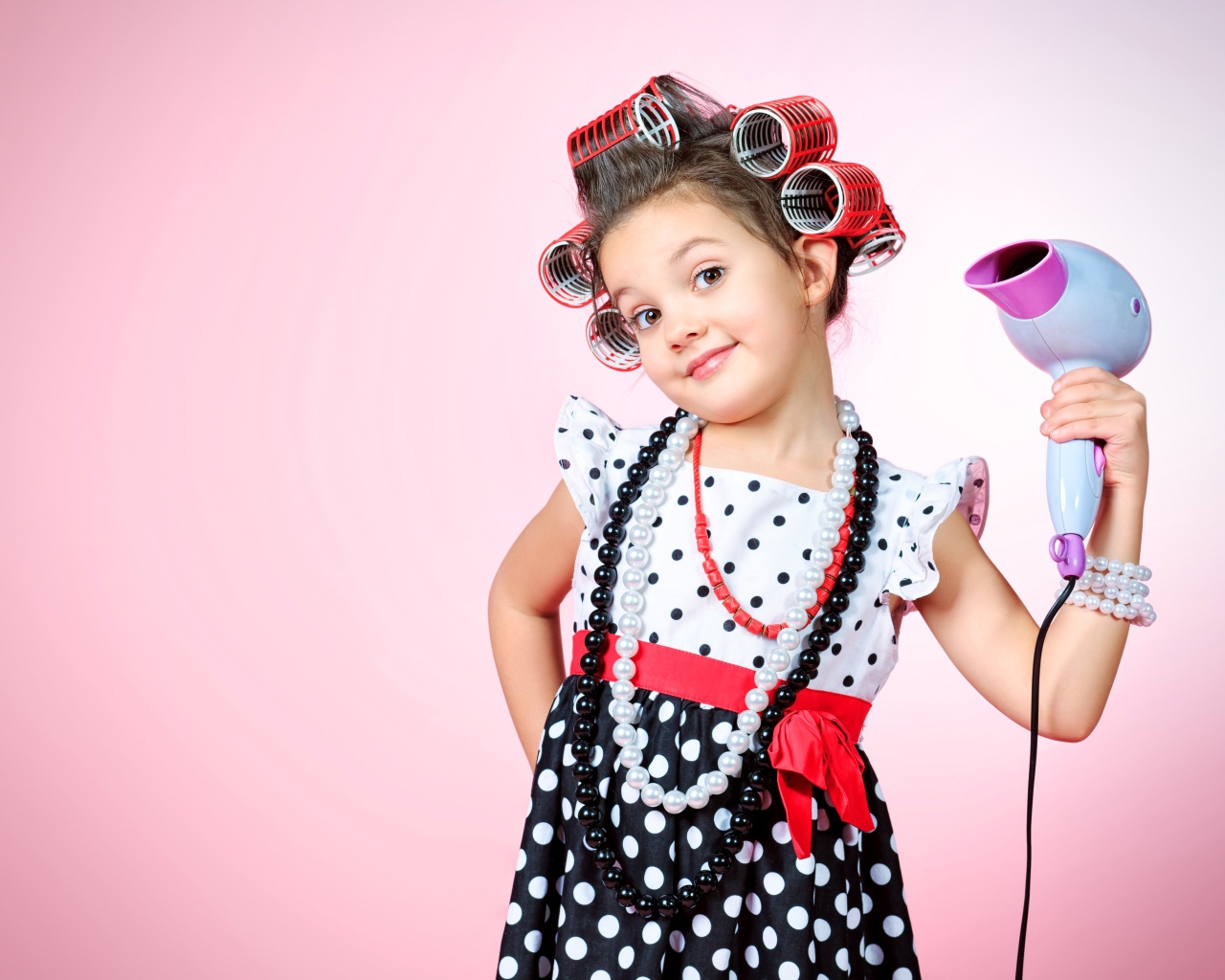 Маленькая девочка с бигудями на голове с феном в руке на розовом фоне