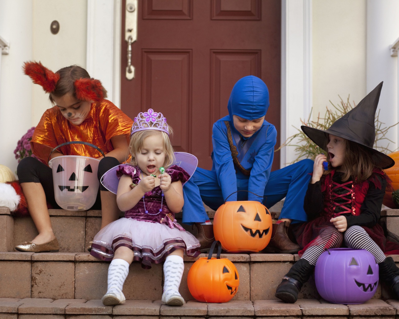 Маленькие дети в костюмах на Хэллоуин сидят на ступеньках со сладостями