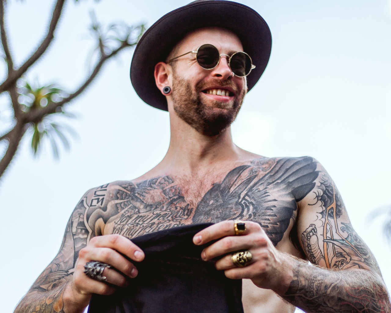 A man with tattoos on his body in a black hat and glasses