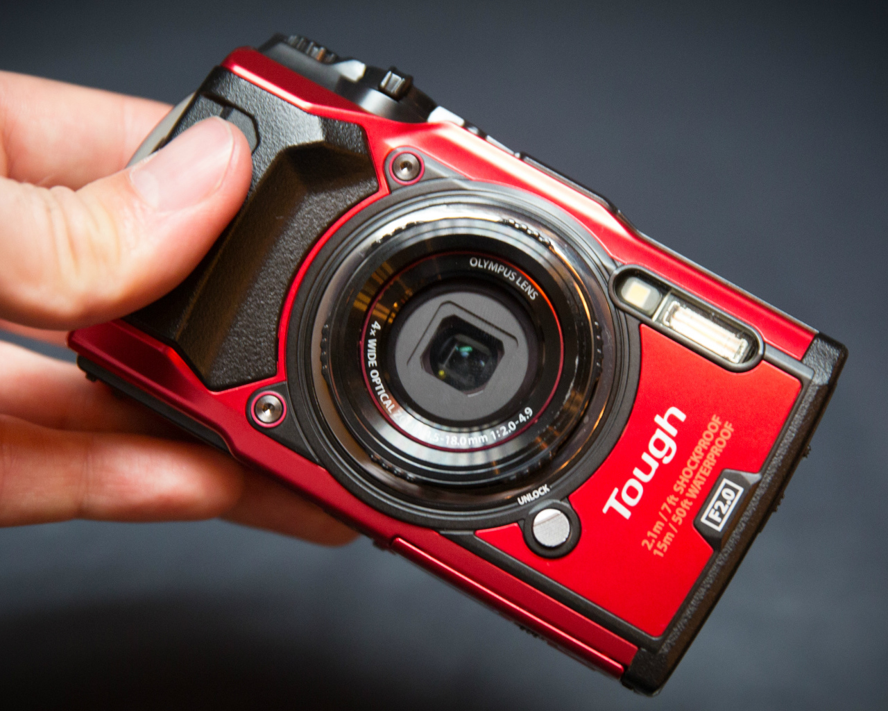 Красный фотоаппарат Olympus Tough TG-5 в руке на сером фоне
