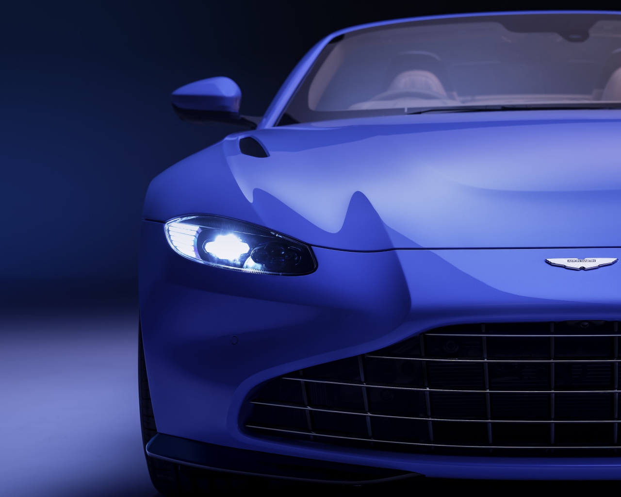 Синий автомобиль Aston Martin Vantage Roadster 2020 года с включенной фарой 