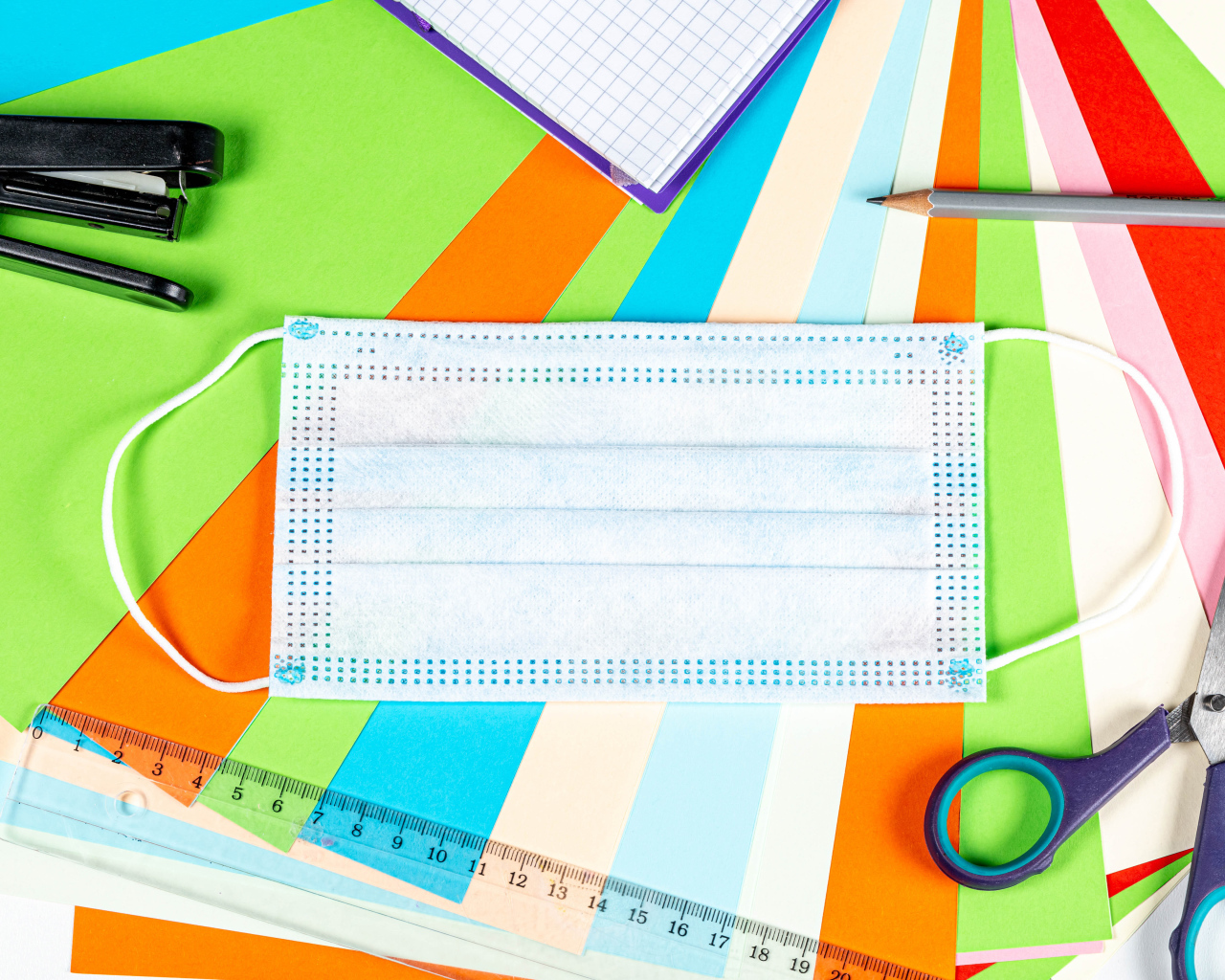 Маска от коронавируса лежит на цветной бумаге с канцелярскими предметами