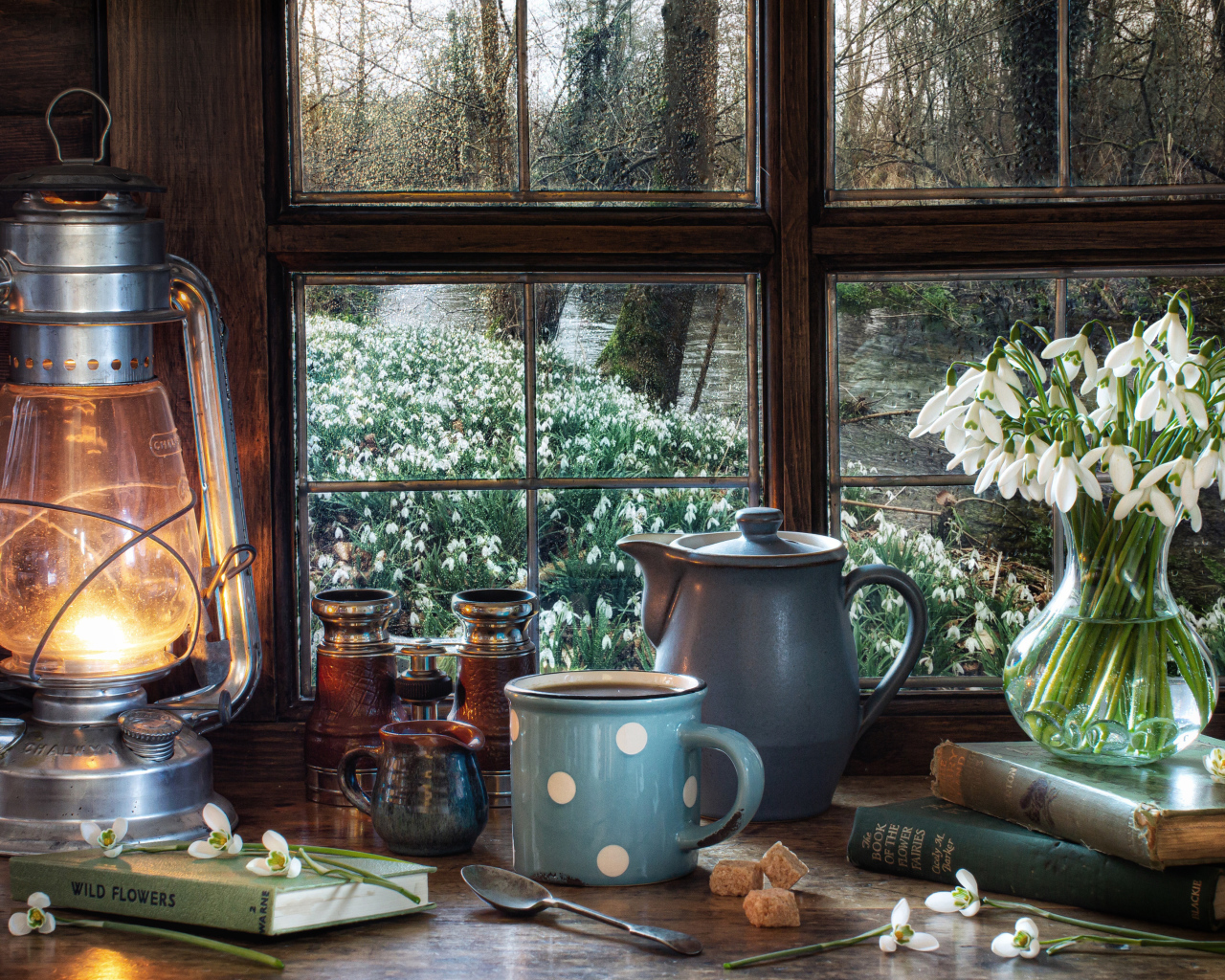 Керосиновая лампа на столе с кружкой, книгами и букетом подснежников у окна