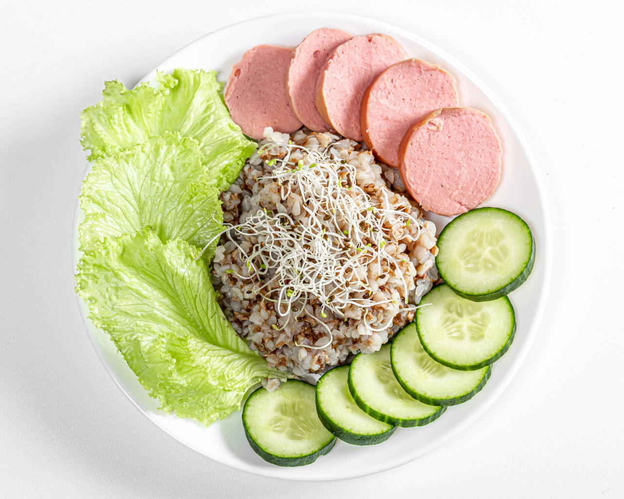 Гречка на тарелке с огурцом, колбасой и листьями салата на белом фоне