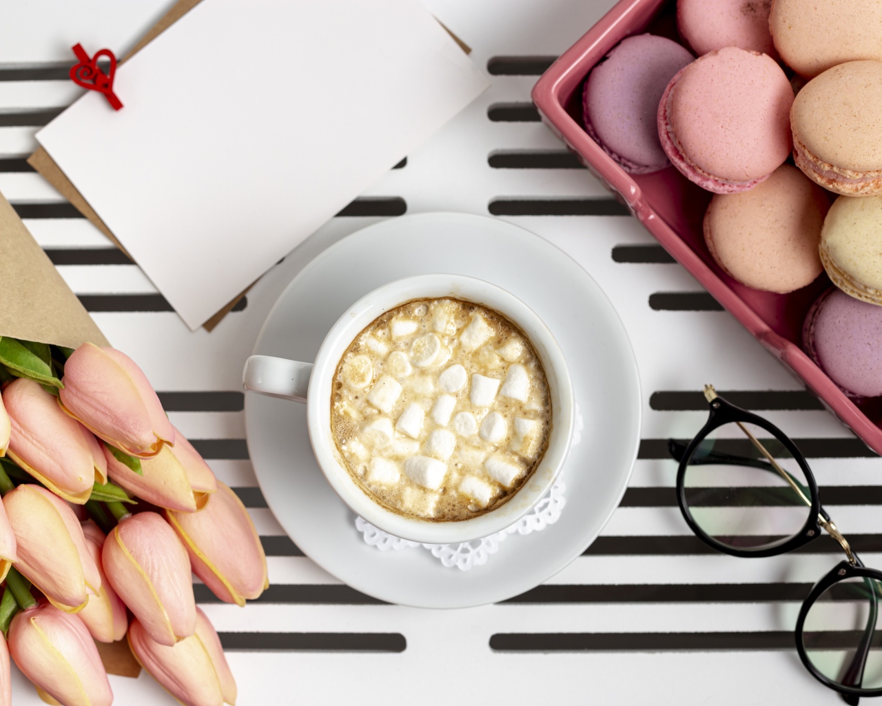 Чашка какао с маршмеллоу на столе с букетом тюльпанов, десертом макарун и очками