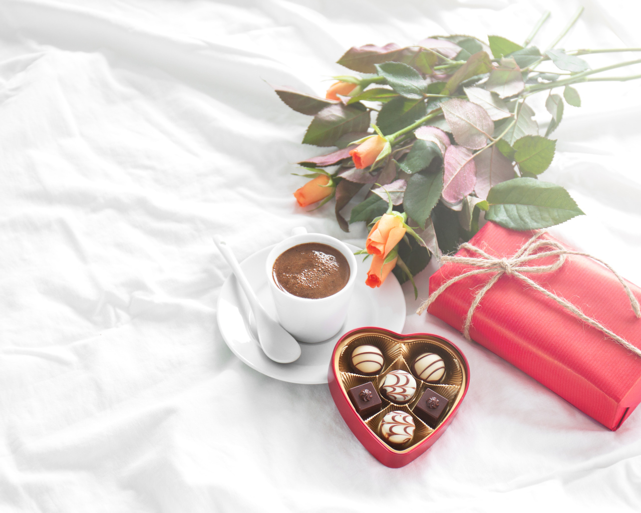Чашка кофе на белой кровати с конфетами, подарком и букетом роз