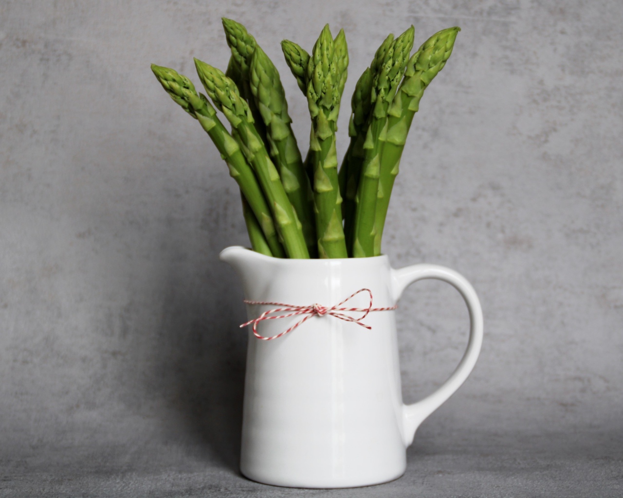 Зеленая спаржа в белой вазе на фоне серой стены