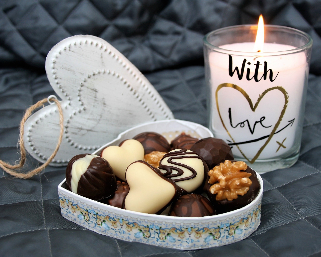 Шоколадные конфеты в коробке в форме сердца на кровати с зажженной свечой 