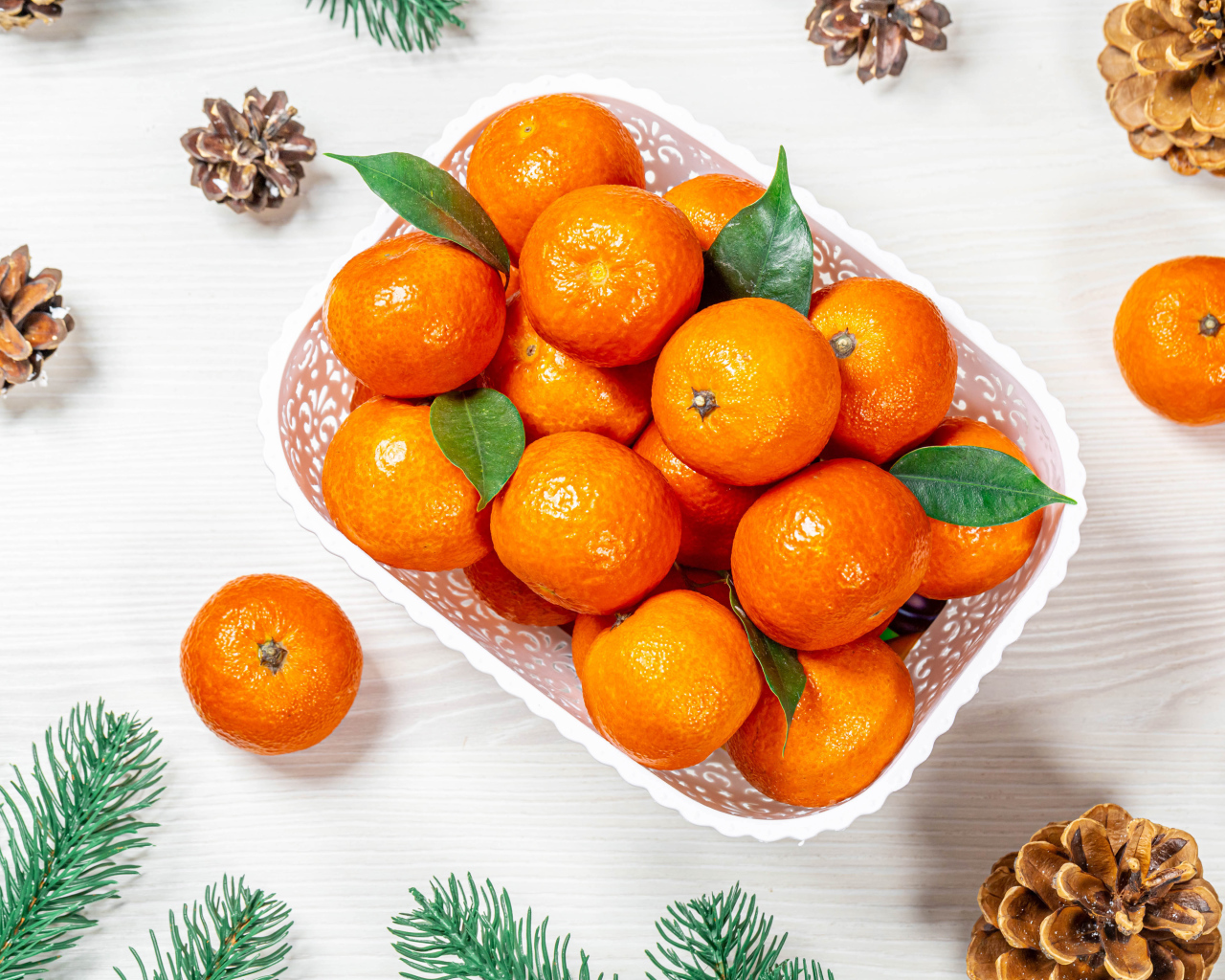 Many ripe tangerines in a white basket on a table with pine cones