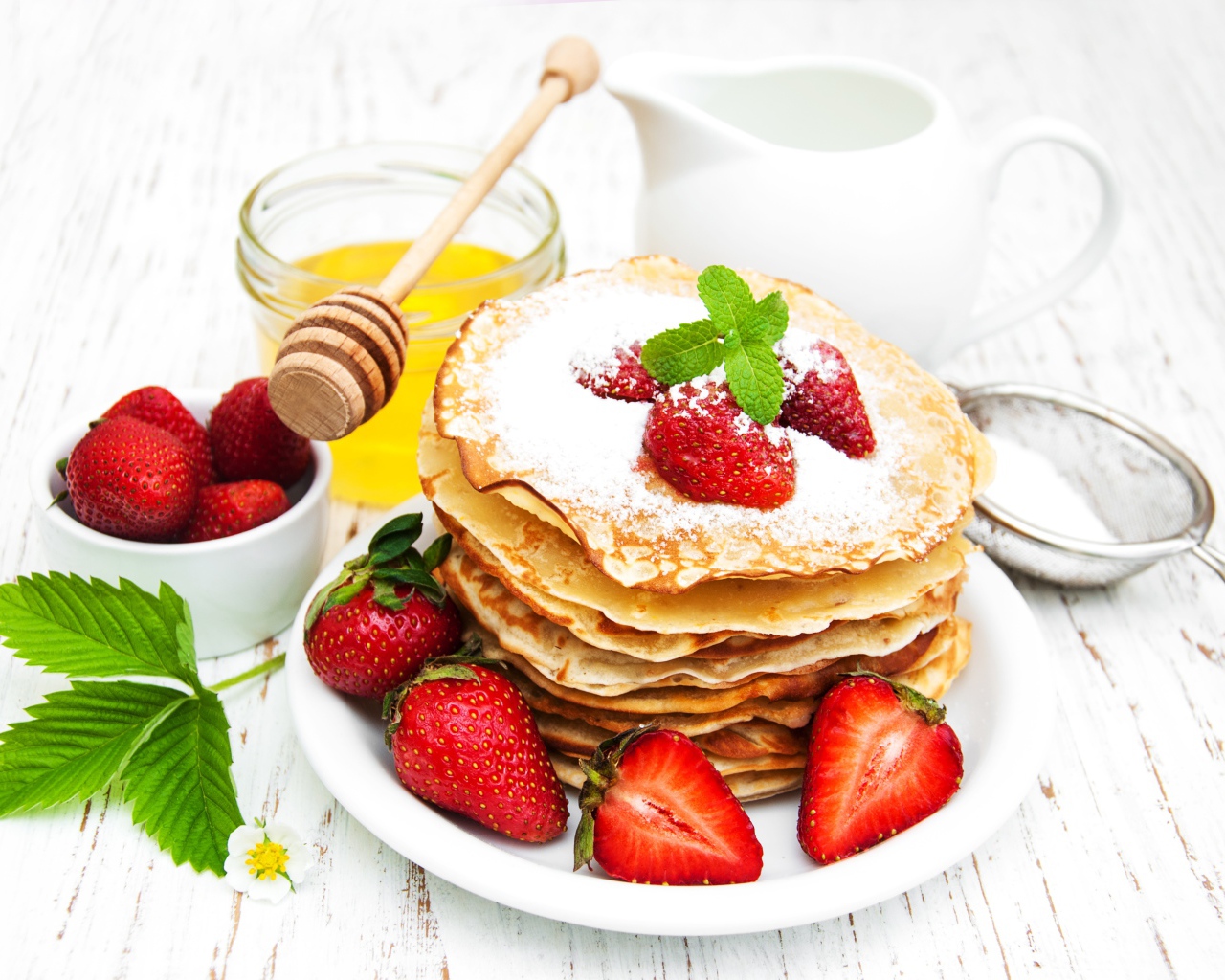 Тонкие блины с сахарной пудрой и ягодами клубники на столе с молоком и медом