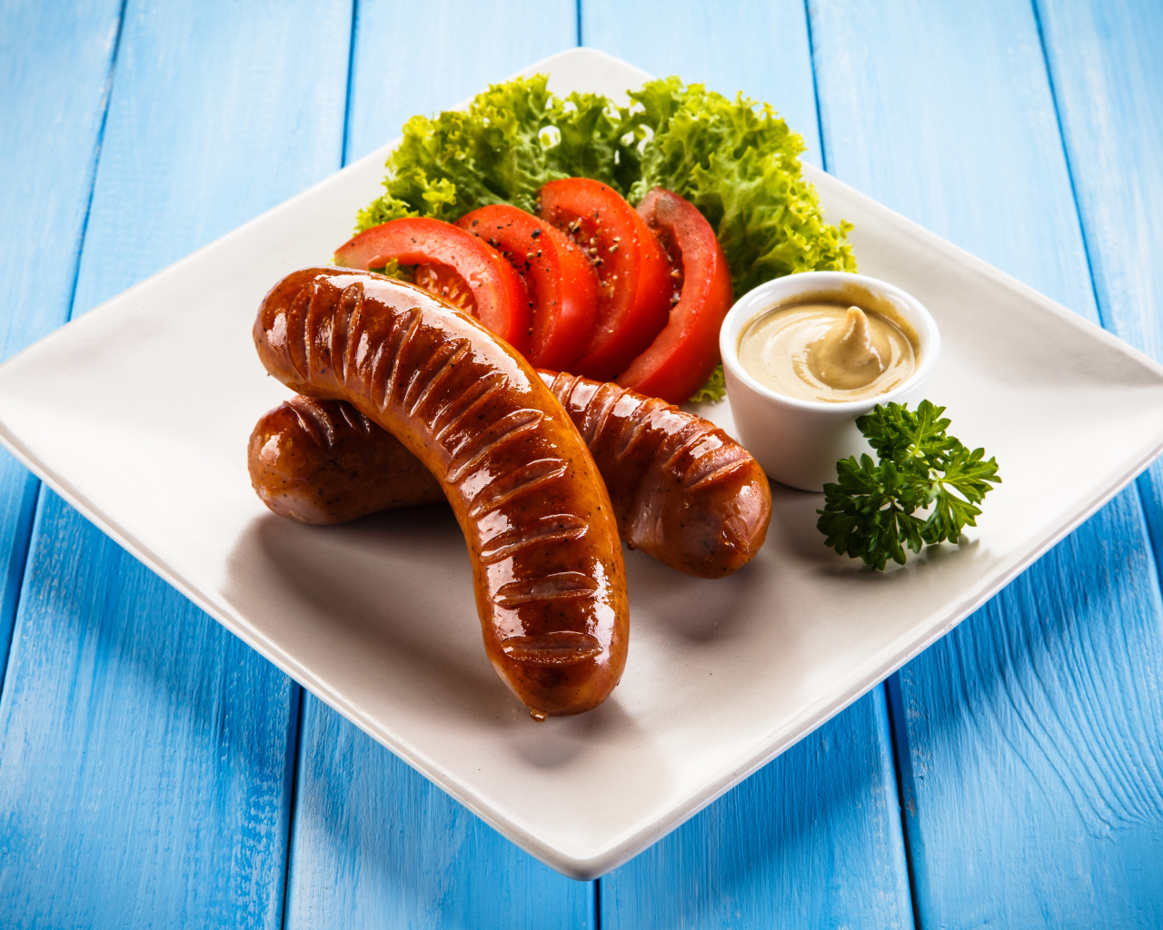 Две жареные колбаски на тарелке с помидорами, горчицей и листьями салата 