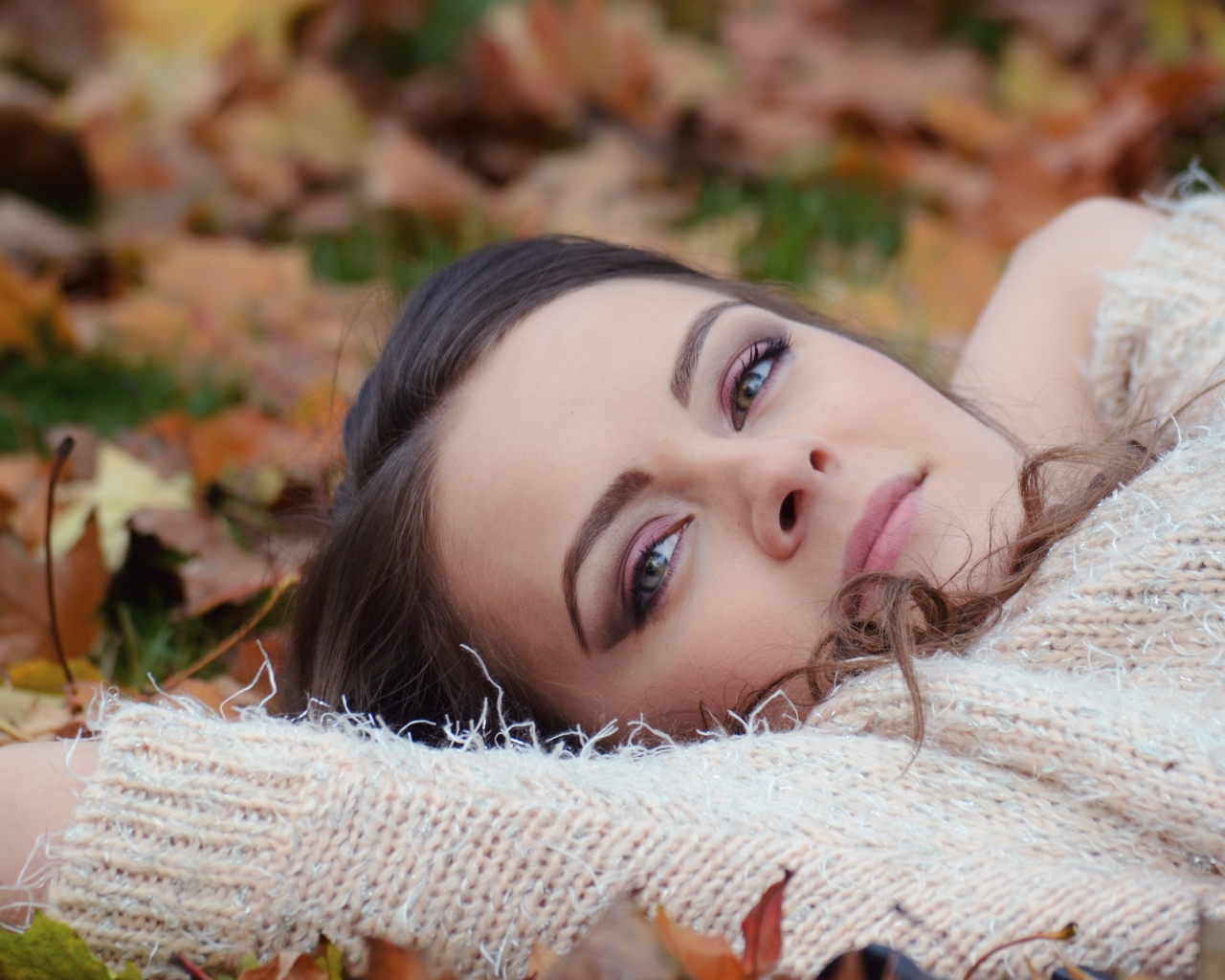 Красивая девушка в теплом свитере лежит на сухой листве 