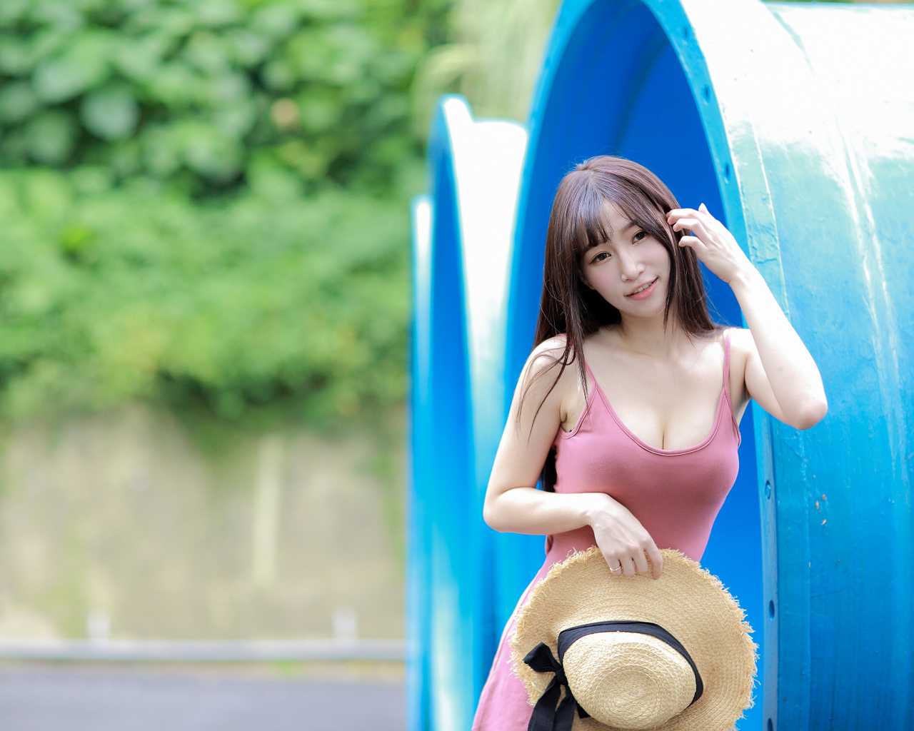 Стройная азиатка в платье со шляпой в руке стоит у синих  труб 