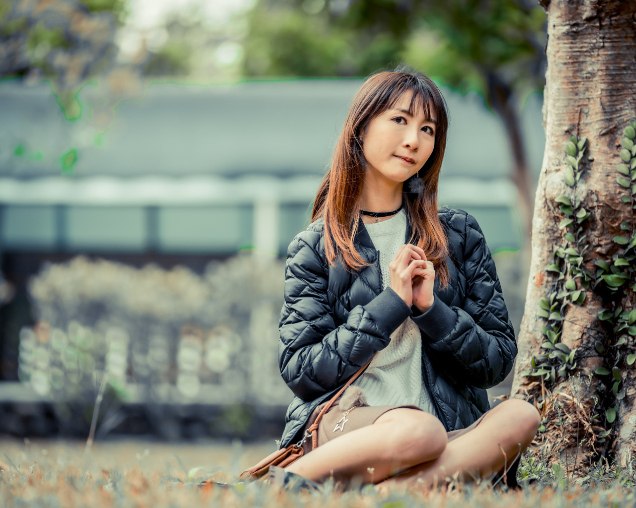 Девушка азиатка в черной куртке сидит под деревом