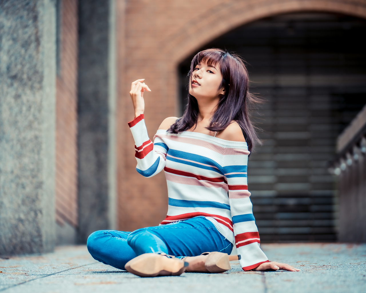 Девушка азиатка в джинсах и полосатом свитере сидит на асфальте
