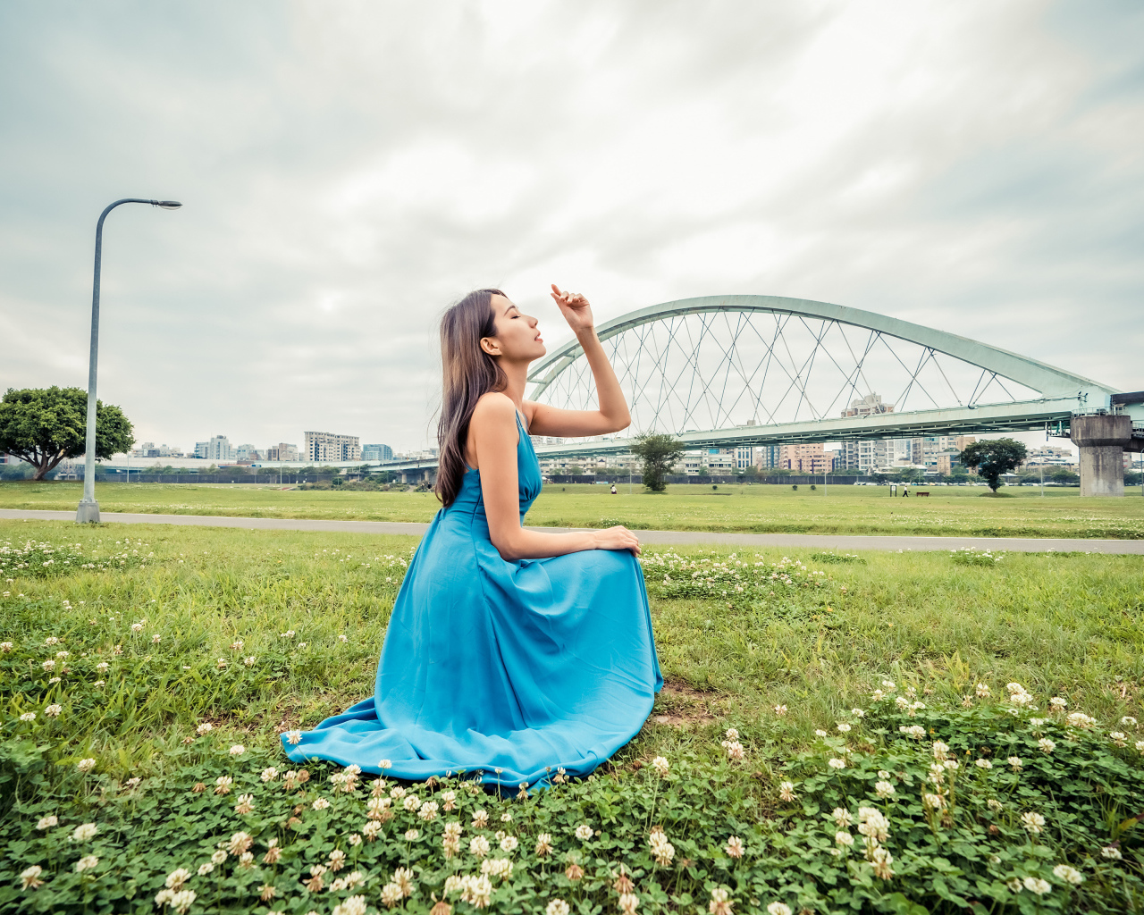 Красивая девушка азиатка в голубом платье  сидит на траве на фоне моста