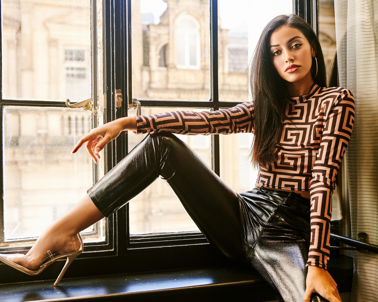 Красивая брюнетка в черных брюках сидит на окне 