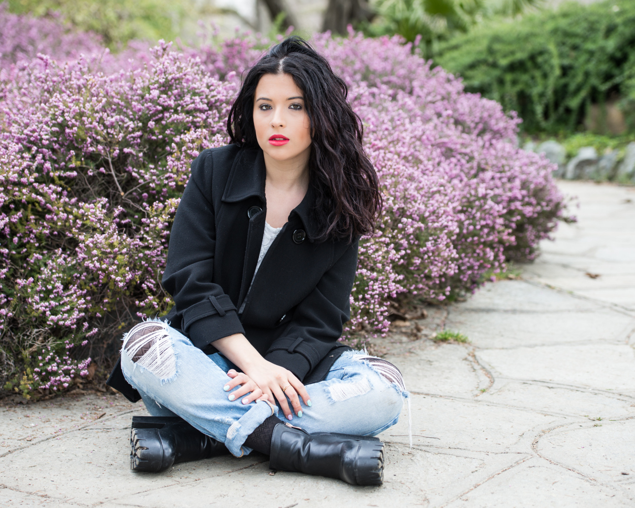 Красивая девушка в черном пальто сидит на земле у кустов