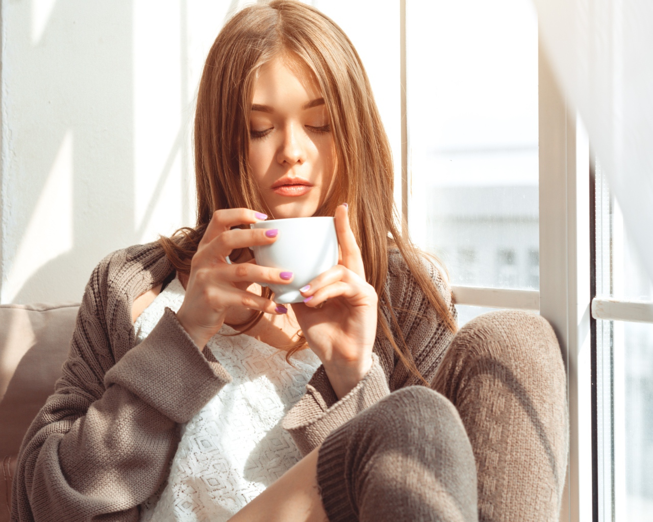 Красивая девушка в свитере пьет кофе у окна