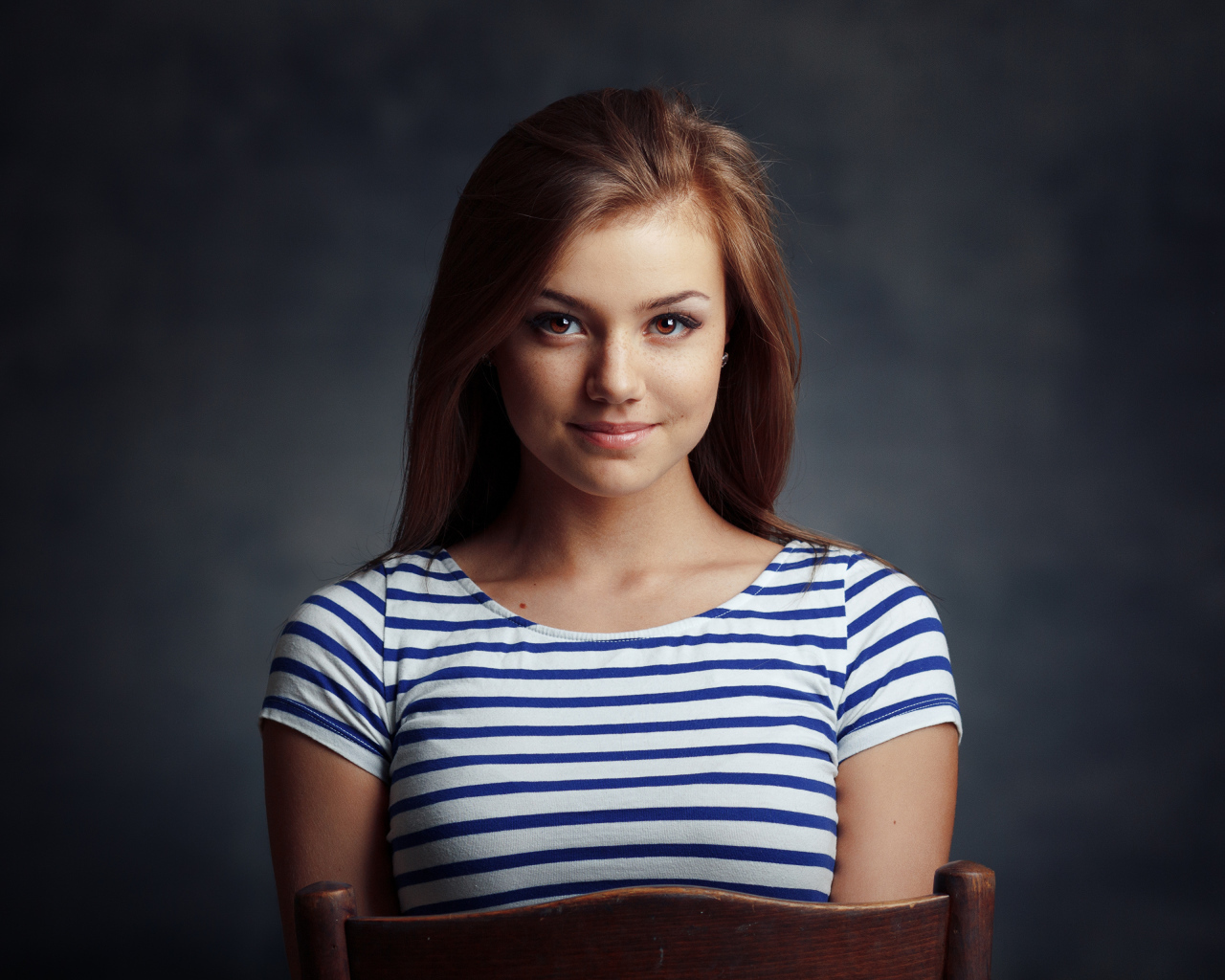 Милая кареглазая девушка в полосатой майке сидит на стуле 