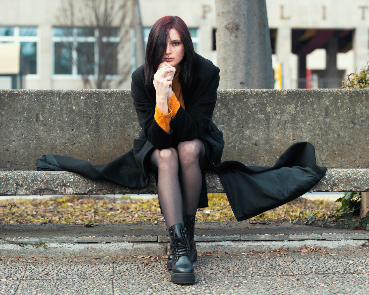 Девушка в черном пальто сидит на бетонной лавке 