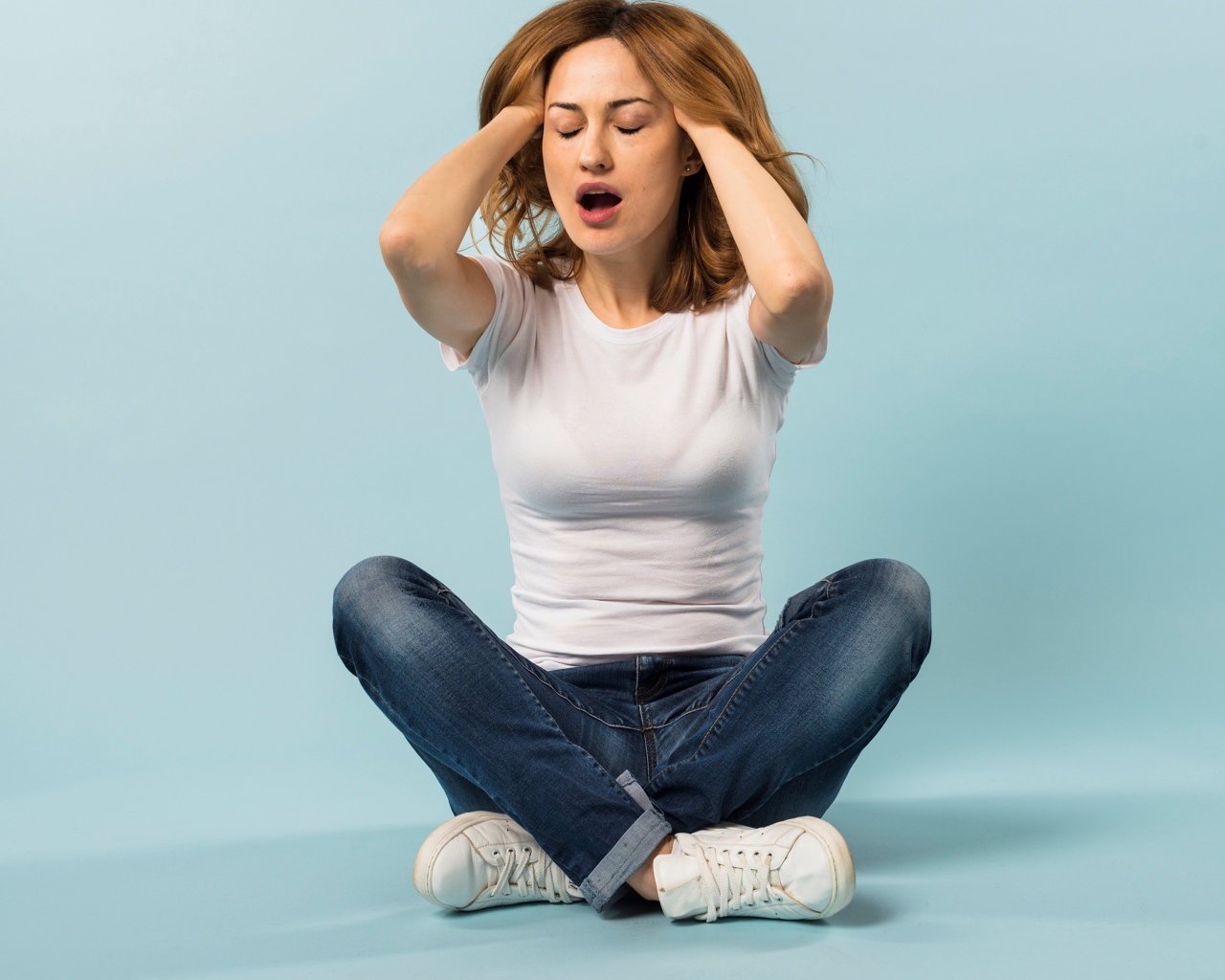 Девушка с руками в волосах сидит в джинсах на голубом фоне