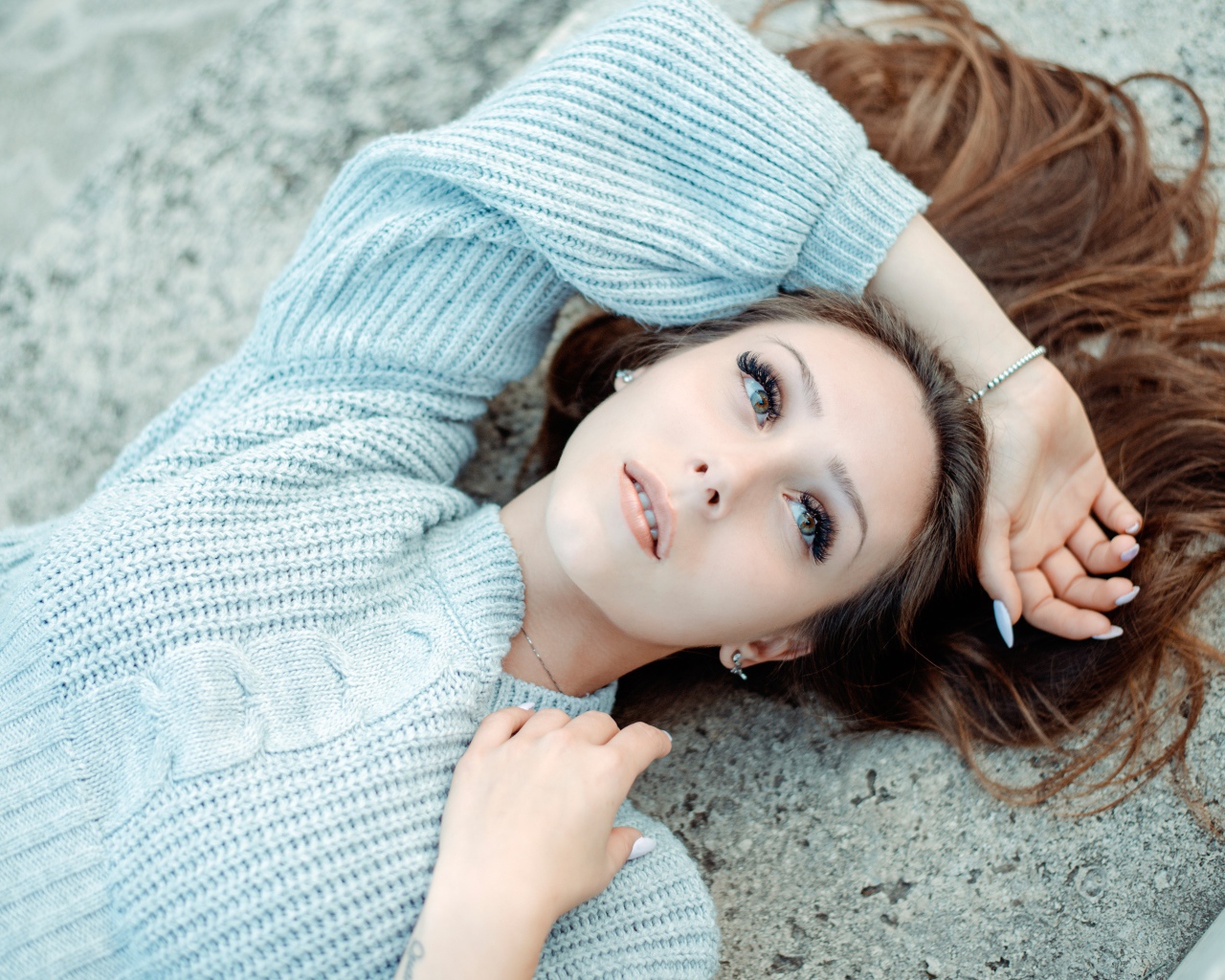 Длинноволосая девушка в голубом свитере лежит на земле 
