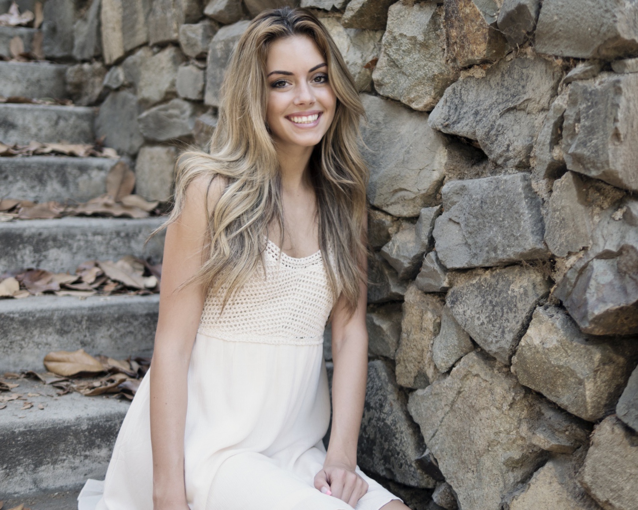 Улыбающаяся  девушка в белом платье стоит на ступеньках у стены