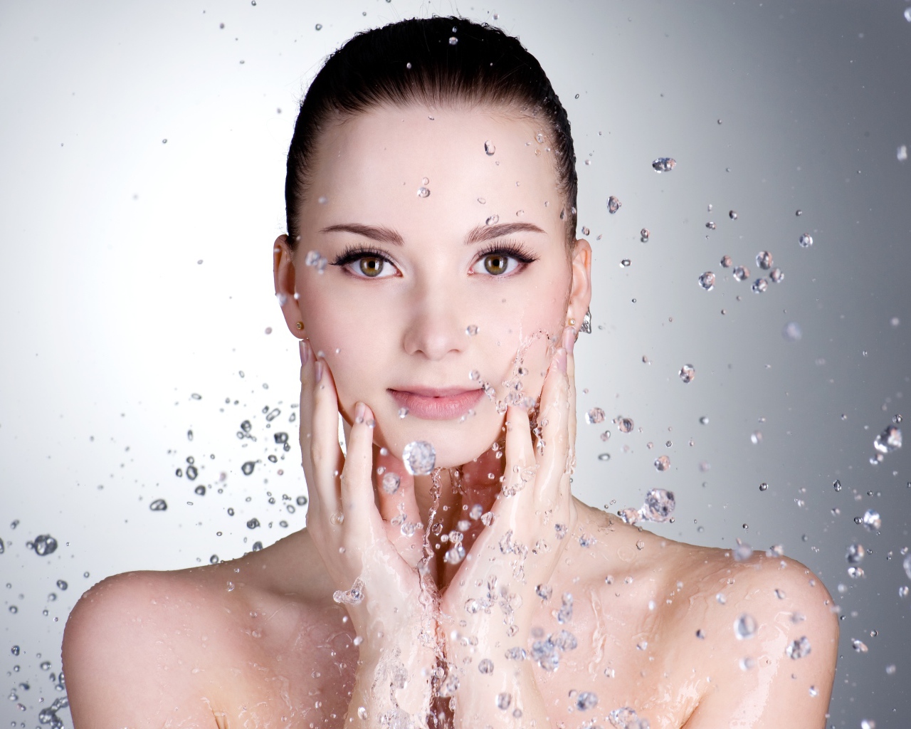 Брызги воды летят на красивое лицо молодой девушки на сером фоне