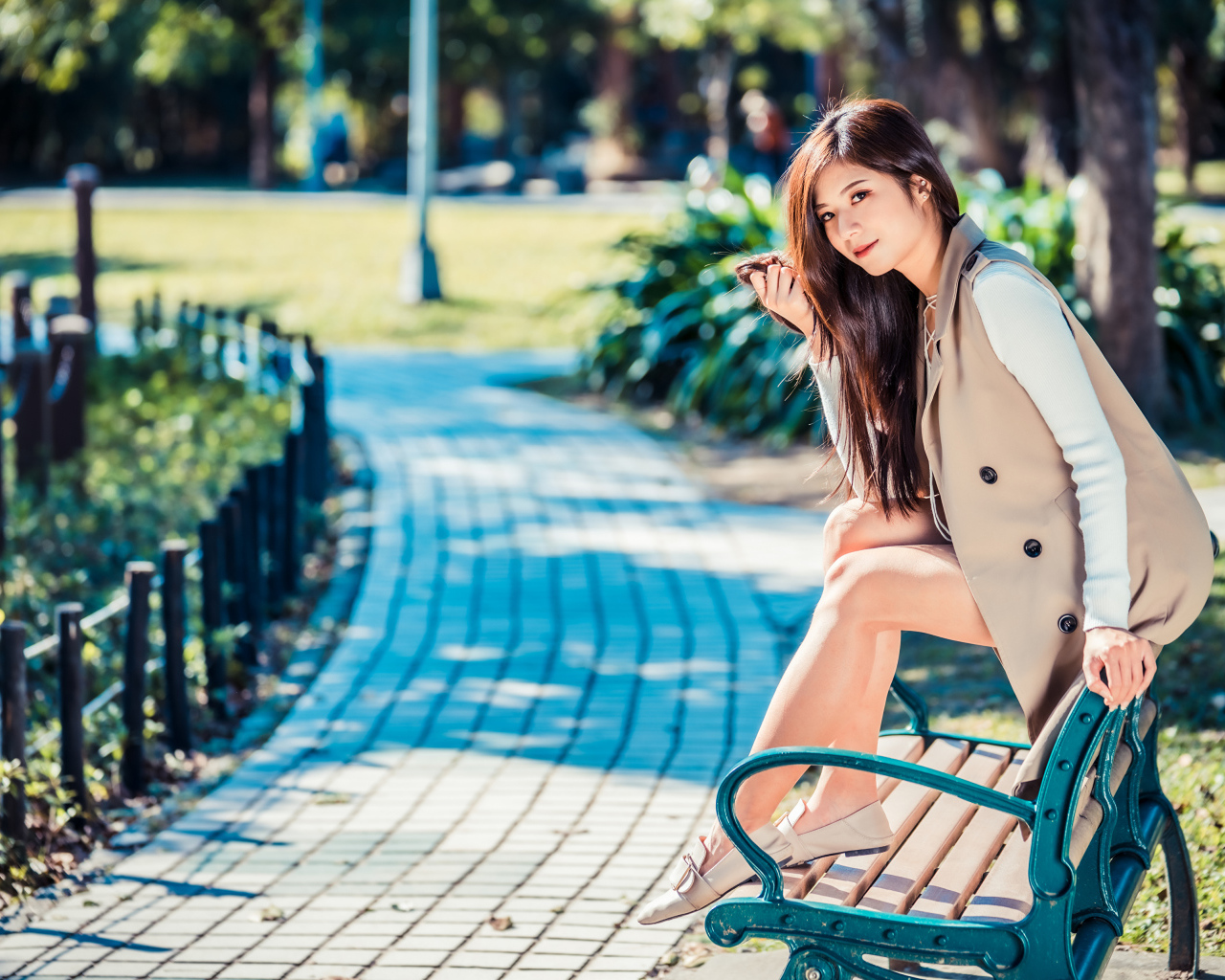 Молодая девушка азиатка сидит в пальто на лавке в парке 