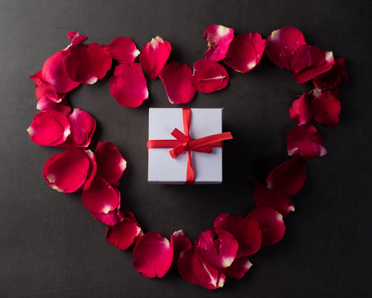 Сердце из красных лепестков розы с подарком на сером фоне